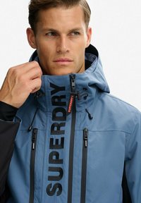 Niebieska wodoodporna kurtka z wysokim kołnierzem, czarnymi akcentami i wyraźnym napisem "SUPERDRY". Posiada regulowane troczki i kieszenie zapinane na zamek.