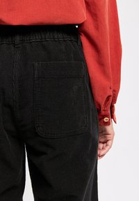 Pantalon en velours noir avec une poche arrière, associé à une chemise rouge à manches longues. Le tissu a une texture côtelée et une taille élastique.