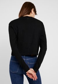 Černá zkrácená košile s dlouhými rukávy, vyrobená z bavlny, s kulatým výstřihem a hladkou strukturou. Zadní strana má minimální branding.