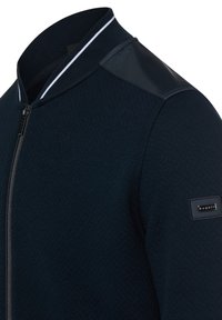 Kurtka bomberka w kolorze granatowym z zamkiem błyskawicznym, wykonana z fakturowanej tkaniny, z prążkowanym kołnierzem i błyszczącymi akcentami na ramionach. Na rękawie znajduje się oznakowana metka.