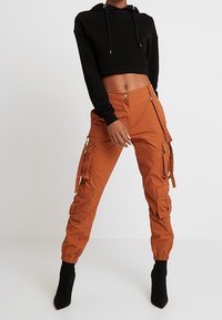 Sudadera negra corta con cordones combinada con pantalones cargo naranjas que tienen bolsillos laterales y tobillos fruncidos. Botas negras de tacón completan el look.