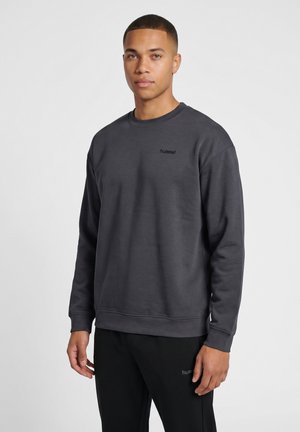 Ung mand stående iført en mørkegrå sweatshirt og sorte bukser med små "hummel" logoer på bryst og lår mod en neutral baggrund.