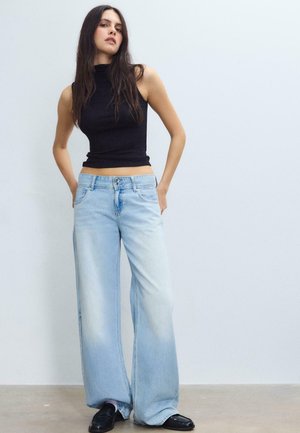 Ung kvinde med langt mørkt hår iført en ærmeløs sort top, lyseblå bukser med vide ben og sorte loafers, stående mod en ensfarvet væg.