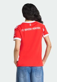Punainen FC Bayern München -paita valkoisilla yksityiskohdilla, jossa on logo ja teksti "MIA SAN MIA" selässä. Lyhyet hihat ja pyöreä pääntie.