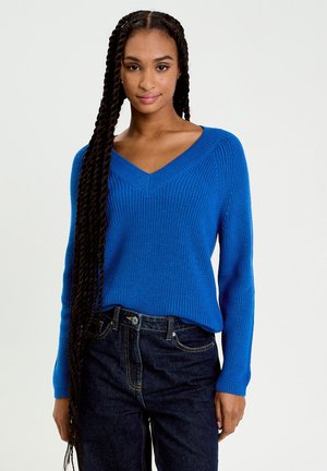 Mujer con el cabello largo y trenzado que lleva un suéter azul brillante de cuello en V y jeans azul oscuro, de pie contra un fondo claro y liso.