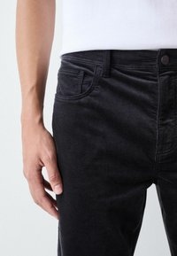 Black corduroy pants with a ribbed texture, featuring side pockets and a button closure, shown on a model's hand resting at the waist.