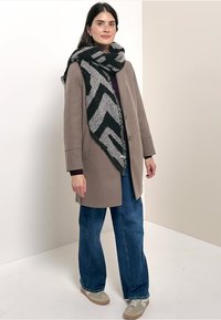 Beige oversized jas, donkerpaarse top, blauwe wijde jeans, grijs-zwart gemarmerde sjaal en lichtbruine sneakers. Eenvoudig, getextureerd ontwerp.