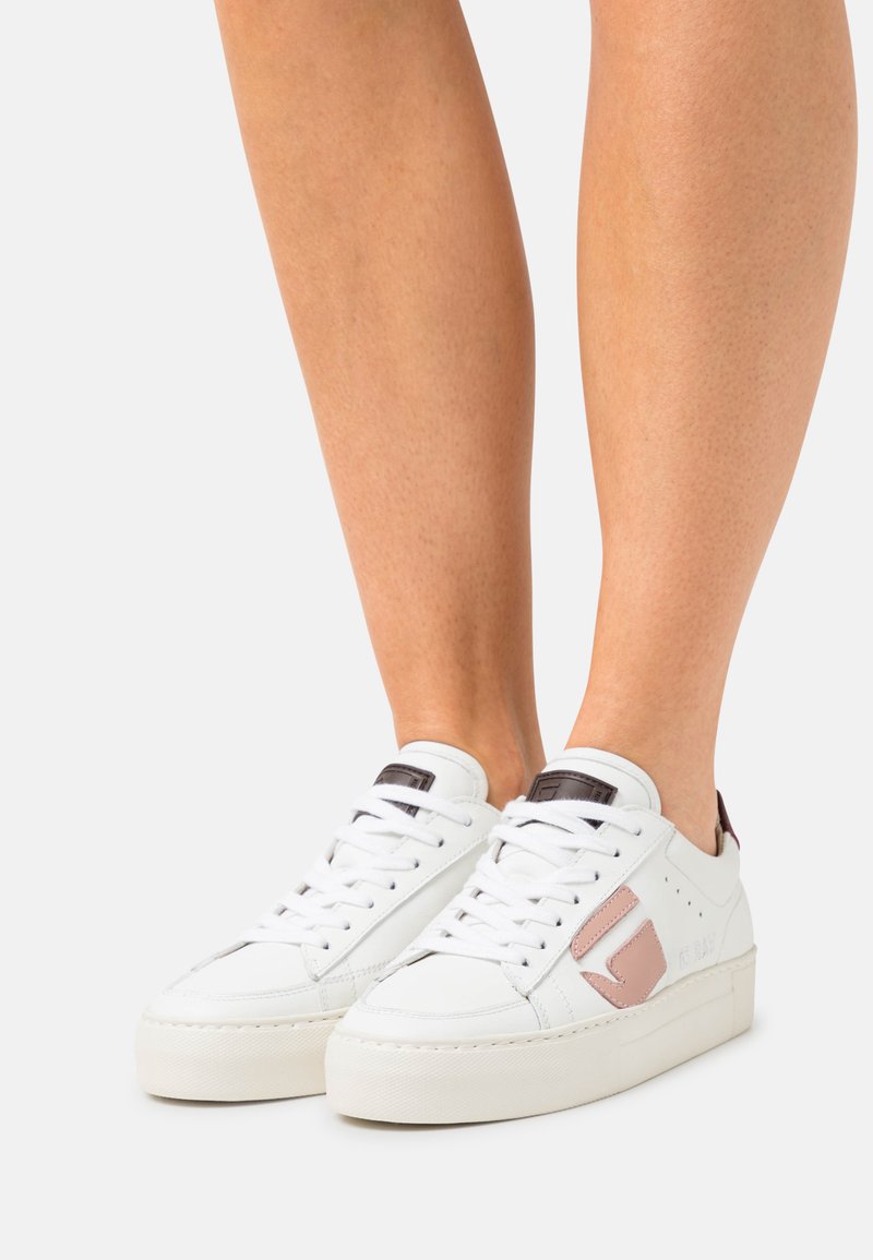 White leather sneakers with a pink accent, flat sole, and thick textured sole. Features laces and tag detail on the tongue.