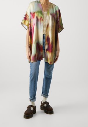 Person trägt ein mehrfarbiges, lockeres Kimono, ein leichtes gehäkeltes Top, hochgekrempelte blaue Jeans, weiße Socken und dunkelbraune Plateauschuhe.