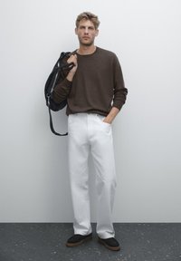 Uomo con camicia a maniche lunghe marrone e pantaloni bianchi, che tiene una borsa nera a tracolla, in piedi contro un muro bianco con una mano in tasca.