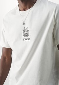 Biała bawełniana koszulka z krótkim rękawem, z wyszywanym czarnym logo przedstawiającym rogate stworzenie oraz napisem "ICEBERG" poniżej. Klasyczny dekolt.
