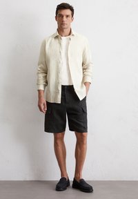Jasno beżowa lniana koszula zapinana na guziki na białym t-shircie, w połączeniu z czarnymi szortami cargo i ciemnymi butami wsuwanymi, na jednolitym tle.