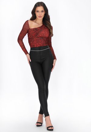 Kobieta stojąca na białym tle, ubrana w czarne spodnie z wysokim stanem, czerwony wzorzysty top na jedno ramię oraz czarne, odkryte szpilki.