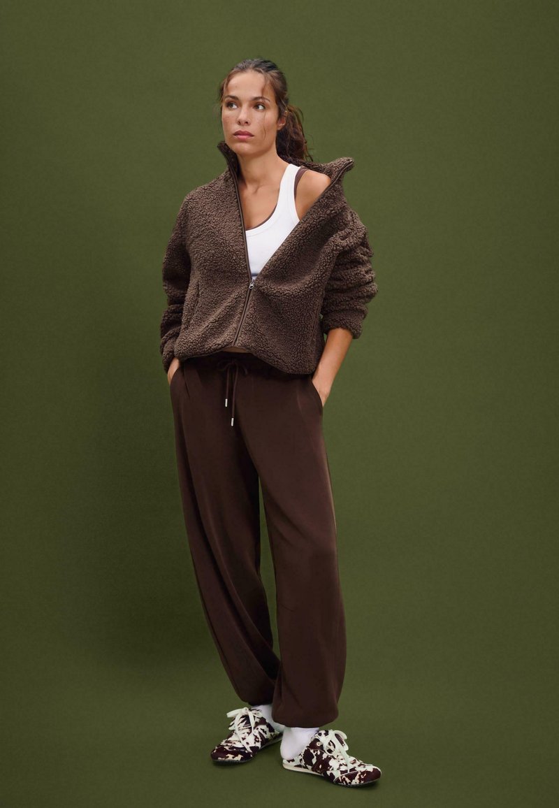 Braune Fleece-Zip-Jacke mit hohem Kragen, kombiniert mit lockeren braunen Hosen und gemusterten Slippern, vor einem grünen Hintergrund.