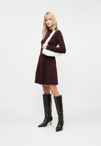 Abito in maglia color bordeaux con maniche lunghe, corpino aderente e gonna svasata. Borsa bianca con tracolla borchiata. Stivali alti in pelle nera con tacco a spillo.