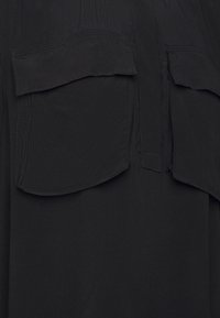 Schwarze Bluse mit zwei großen Brusttaschen, glatter Textur, Knopfleiste und dezenten Falten. Der Stoff wirkt leicht und atmungsaktiv.