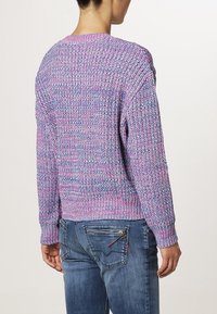 Maglione lavorato a maglia viola con un motivo texturizzato, scollatura rotonda e polsini a costine. Indossato con jeans blu dotati di una tasca posteriore con zip.