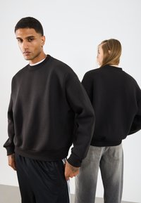 Svart oversized genser med rund hals, ribbede mansjeter og nederkant. Stoffet har en glatt tekstur, vist på to modeller fra front og bak.