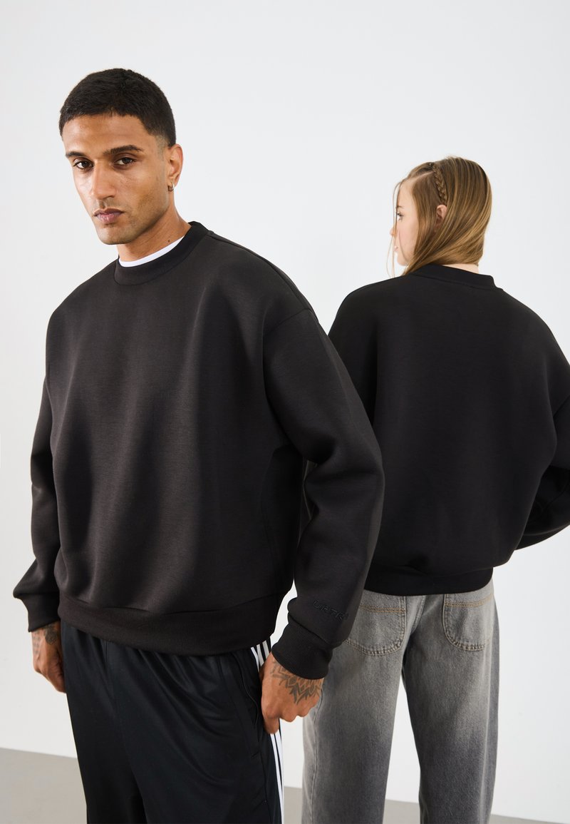 Svart oversized genser med rund hals, ribbede mansjeter og nederkant. Stoffet har en glatt tekstur, vist på to modeller fra front og bak.