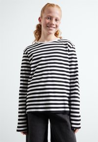 Chemise à rayures noires et blanches à manches longues avec une coupe décontractée et un col rond, associée à un pantalon gris foncé. Le tissu semble lisse.