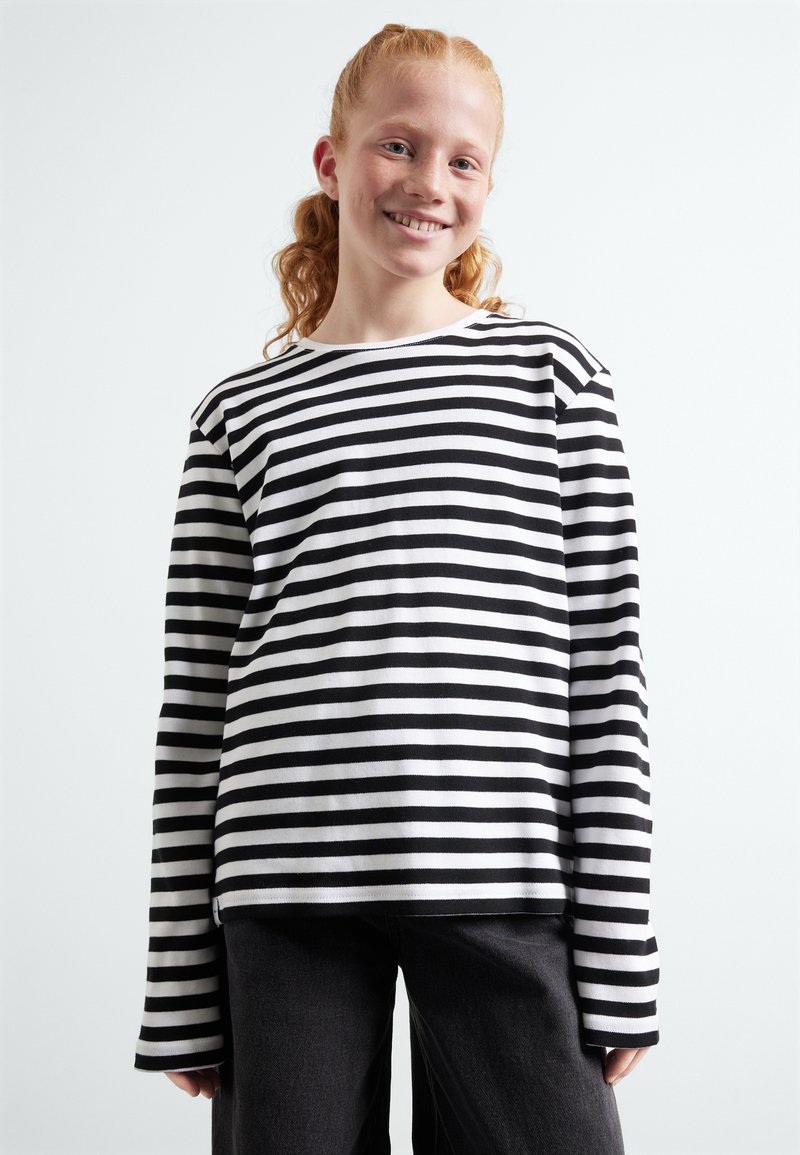 Chemise à rayures noires et blanches à manches longues avec une coupe décontractée et un col rond, associée à un pantalon gris foncé. Le tissu semble lisse.