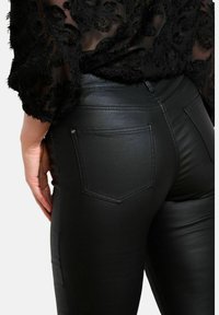 Zwarte leren leggings met een gladde textuur, voorzien van platte zakken en verhoogde stiknaaddetails aan de achterkant. Gestyled met een doorzichtige zwarte top.