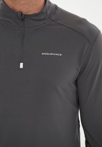 Donkergrijze langwerpige trui met een kwart rits en een pasvorm ontwerp. Kenmerkt zich door een klein "ENDURANCE" logo in het wit op de borst. Glad textuur.