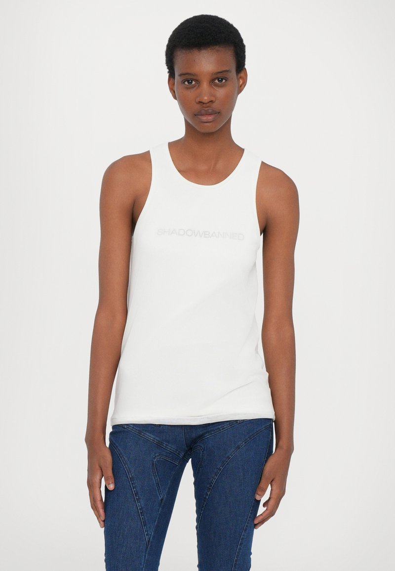 Top sin mangas blanco con la inscripción "SHADOWBANNED" en gris claro, con un escote redondo. Combinado con jeans azul oscuro.