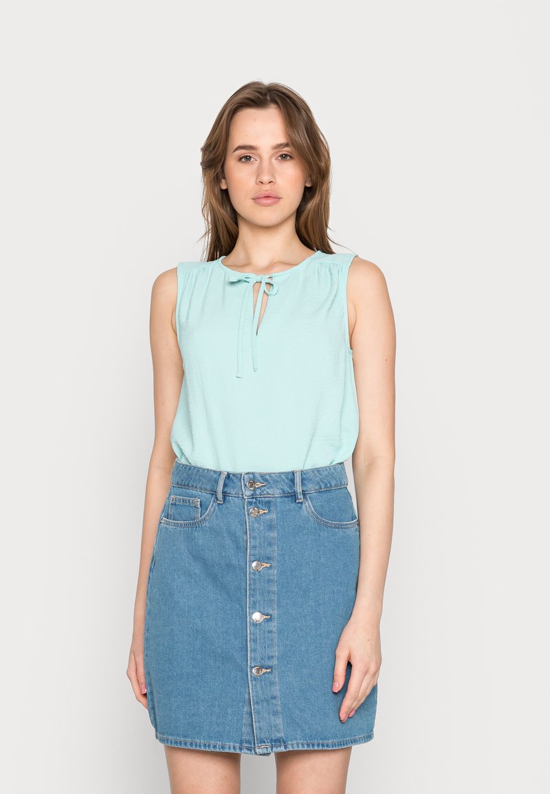 Blusa sem mangas, azul claro, com um laço no decote, combinada com uma saia de ganga com botões na frente e uma cintura ajustada.