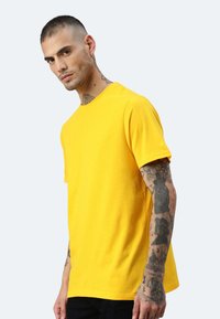 Gelbes T-Shirt mit kurzen Ärmeln und rundem Ausschnitt. Weiches Baumwollmaterial, lockere Passform, mit Tätowierungen auf dem Arm vor einem hellen Hintergrund.