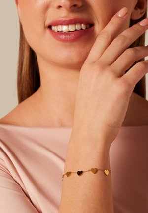 Close-up van een vrouw die lacht, gekleed in een roze top en een gouden armband met kleine hartvormige bedeltjes om haar pols.