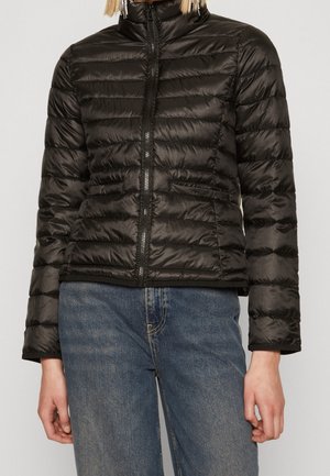 Person iført en sort quiltet dunjakke med lynlås foran og blå jeans, stående mod en ensfarvet lys baggrund.