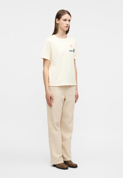 Νεαρή γυναίκα όρθια, φοράει ένα κρεμ κοντομάνικο μπλουζάκι με μπαλώματα, μπεζ παντελόνι φαρδύ στη μπότα και καφέ παπούτσια με χαμηλό τακούνι σε λευκό φόντο.