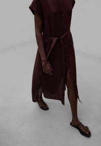 Tumšas ādas cilvēks staigā, valkājot tumši brūnu pogātu kleitu ar jostas saitīti un melnas plakanas sandales uz gaiši pelēkas grīdas.