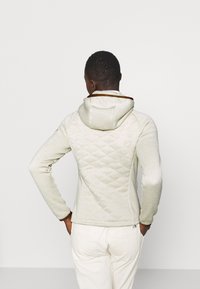 Uomo in piedi con le mani in tasca, che indossa un hoodie trapuntato di un leggero beige e pantaloni bianchi, rivolto di spalle su uno sfondo neutro.