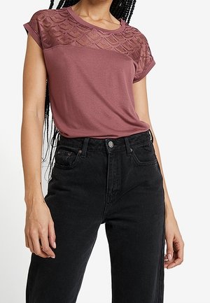 Kvinde iført en støvet lilla kortærmet top med blonde detaljer ved skuldrene og sort, højtaljede jeans, stående mod en hvid baggrund.