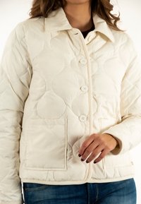 Giacca trapuntata color crema con colletto, cinque bottoni e due tasche frontali con dettagli di cucitura visibili.