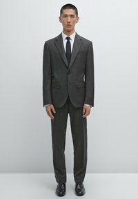 Grauer Anzug mit Spitzrevers, zwei Fronttaschen und einem Ein-Knopf-Verschluss, kombiniert mit einem weißen Hemd und einer marineblauen Krawatte. Schwarze Schuhe runden den Look ab.
