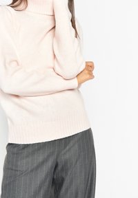 Un pull col roulé rose clair en matière tricotée douce avec des poignets et un ourlet côtelés, associé à un pantalon gris à rayures.
