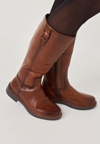 Stivaletti al ginocchio in pelle marrone con cerniera laterale, caratterizzati da una fibbia decorativa e una superficie liscia, su sfondo bianco.