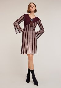 Burgundy ja raidallinen mekko, jossa on pitkät, poimutetut hihat, syvä pääntie ja tupsukoriste. Rakennekerros voi olla karheaa, mikä luo kontrastin sileiden alueiden kanssa.