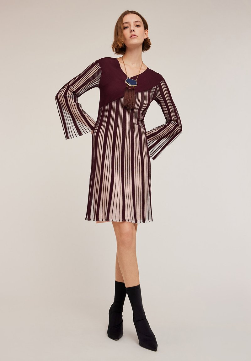 Burgundy ja raidallinen mekko, jossa on pitkät, poimutetut hihat, syvä pääntie ja tupsukoriste. Rakennekerros voi olla karheaa, mikä luo kontrastin sileiden alueiden kanssa.