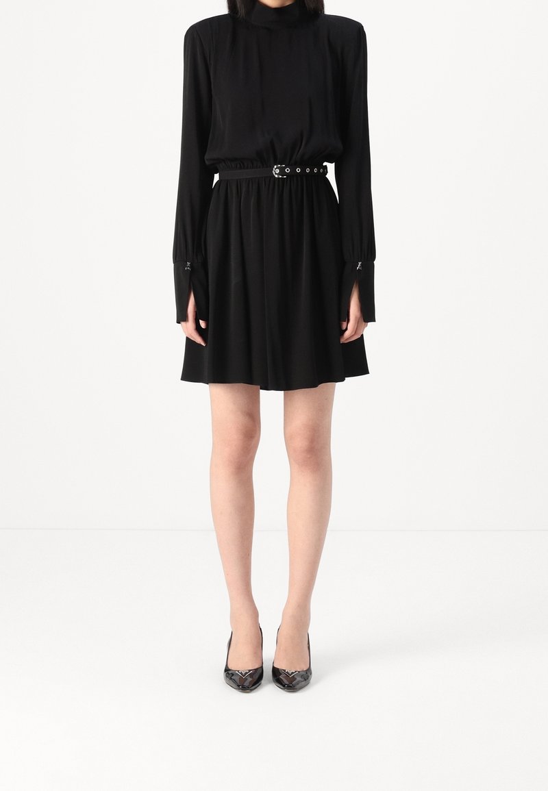 Femme portant une robe noire à manches longues avec une ceinture à la taille et des talons aiguilles noirs pointus, debout devant un fond blanc.