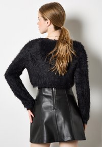 Vrouw met een lange paardenstaart, gekleed in een zwarte fuzzy trui en een hoge, zwarte leren rok met een zichtbare ritssluiting aan de achterkant, staande tegen een witte muur.