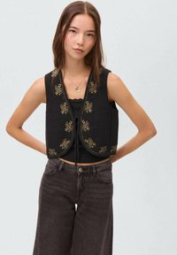 Črn prešit brezrokavnik z zlatim cvetličnim vezenjem, zasnova z odprtim sprednjim delom, nošen preko črne majice in v kombinaciji z visokim pasom, temnimi kavbojkami.
