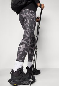 Leggings i sort og grå abstrakt mønster, parret med sorte vandrestøvler og trekkingstænger, med et lille logo på benet.