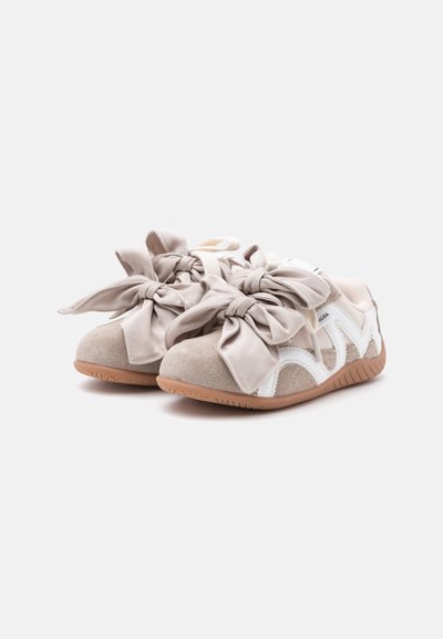 Paire de baskets beige et blanches avec de grands nœuds en ruban de satin comme lacets et semelles en caoutchouc sur fond blanc.