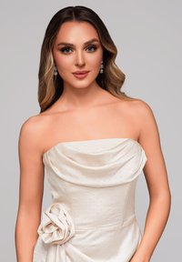 Strapless jurk van crèmekleurige stof met een subtiel stippenpatroon, voorzien van een gedrapeerde halslijn en een bloemenaccent aan de zijkant.
