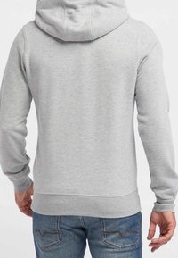Grauer Kapuzenpullover mit Kordelzug, aus weichem Material. Verfügt über eine Kängurutasche und einen gerippten Saum. Kombiniert mit blauen Jeans.