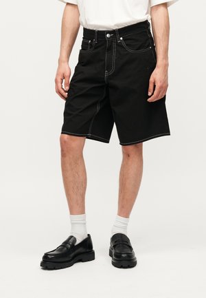 Homme portant un short noir longueur genou avec coutures blanches contrastantes, chaussettes blanches et mocassins en cuir noirs, debout avec les mains détendues.